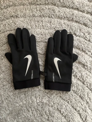 Svarta Nike Therma-Fit handskar - Snygga svarta handskar från Nike med Therma-Fit-teknologi och stor vit logga på ovansidan. Perfekta för kyliga dagar och har en stilren, sportig look. Mjukt och flexibelt material som sitter skönt på handen.