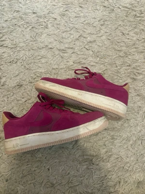Vinröda Nike Air Force 1 sneakers - Säljer ett par vinröda/lila Nike Air Force 1 sneakers i mocka med beige detalj på hälen och vit sula. Klassisk låg modell med snörning och perforeringar på tån. Swoosh-logga på sidan och AIR-text på sulan. Perfekta för dig som vill sticka ut med färg. Pris kan diskuteras.