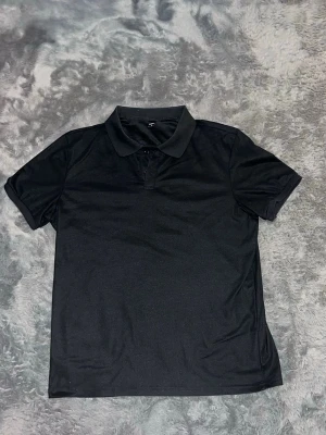 Svart pikétröja med krage - En stilren svart pikétröja med klassisk krage och korta ärmar. Tröjan har knappar vid halsen och är tillverkad i ett mjukt material som känns bekvämt mot huden. Längd 71cm