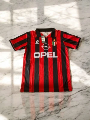 AC Milan retro fotbollströja Maldini - Klassisk AC Milan retro fotbollströja från Lotto med Maldini och nummer 3 på ryggen. Röd och svart randig design, kort ärm, krage och tryckt Opel-logga på bröstet. Perfekt för dig som älskar vintage fotbollströjor och italiensk fotboll.