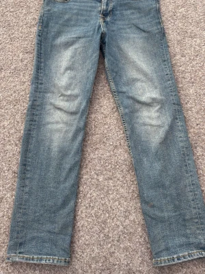 Blå raka jeans - Ett par klassiska blå jeans med raka ben och diskreta slitningar på framsidan. Jeansen har fem fickor och normal midja. Materialet är denim i bomull och färgen är tvättad blå med ljusare partier på lår och knän.