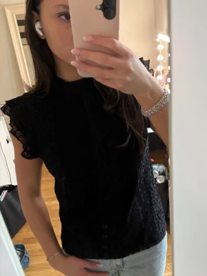 Svart spetsblus med volangärm - Säljer en svart blus i spets med volangdetaljer vid ärmarna. Blusen har hög hals och är ärmlös med en feminin och elegant look. Perfekt att styla med jeans eller kjol för en trendig outfit.