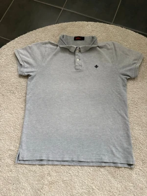 Ljusgrå pikétröja från Morris - Klassisk ljusgrå pikétröja från Morris med korta ärmar och krage. Tröjan har två knappar framtill och en liten mörkblå broderad detalj på bröstet. Perfekt till jeans eller shorts för en enkel och stilren look.