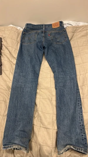 Levi's 501 blå - Klassiska blå 501 från Levis. Väl använda och har en del egengjorda slitningar. Ända defekten är att läderlappen på baksidan har spruckit men inget som påverkar utseendet utan ger bara en nice touch. Måtten är 27cm i midjan och 102 i längd. Storlek är 25/32 på tagen