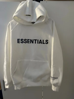 Essentials - Säljer en vit hoodie från Essentials med svart text på bröstet och mindre logga på ärmen. Hoodien har stor huva och magficka framtill. Tillverkad i mjukt bomullsmaterial och har en avslappnad passform. Perfekt för en clean och stilren streetwear-look.