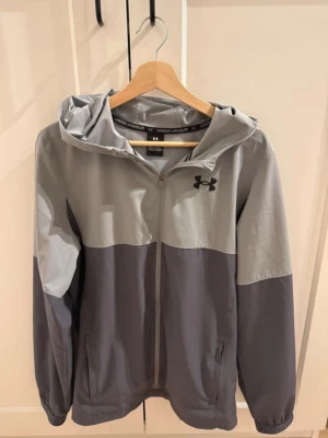 Grå vindjacka från Under Armour - Snygg tvåfärgad grå vindjacka från Under Armour med huva och dragkedja framtill. Jackan har elastiska muddar vid ärmsluten och två sidofickor. Tillverkad i lätt och vindtätt material, perfekt för blåsiga dagar. Under Armour-logga på bröstet.