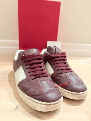 Vinröda Valentino sneakers i läder - Snygga sneakers från Valentino i vinrött och vitt läder. Klassisk låg modell med vita detaljer på sidorna och matchande vinröda skosnören. Det finns defekter i hälarna där lädret har nötts bort men är inget som varken syns eller känns. Tyvärr är även flertalet bitar av med annars är skorna i bra skick. Box samt kuvert med kort  ingår! Fundera inte att fråga om du har några frågor😊