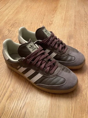 Adidas Samba - Ett par Adidas Samba sneakers i brunt läder med gråa och crémevita mockadetaljer. Skorna har bruna skosnören och gummisula. De är knappt använda och är i princip nya. Äkta, inga defekter och strl 42.