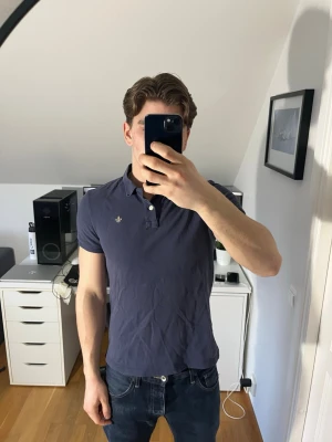 Mörkblå pikétröja från Morris - Säljer en mörkblå pikétröja från Morris med klassisk krage och knappar framtill. Tröjan har en broderad logga på bröstet. Hör gärna av dig vid frågor!