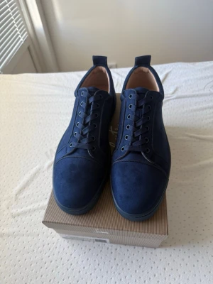 Marinblå sneakers i mocka från Christian Loubutin - Snygga marinblå sneakers i mocka med matchande snören och klassisk låg profil. Skorna har en röd sula som ger en cool kontrast och stilren look. Perfekta för dig som vill ha en clean och modern sneaker med premiumkänsla.