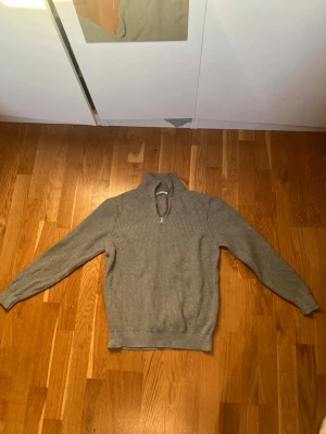 Grå stickad halvzip från Zara - Snygg grå stickad tröja från Zara med halvzip och hög krage. Tröjan har ribbstickade muddar vid ärmslut och nederkant, perfekt för lager på lager. Tillverkad i Bulgarien och har en klassisk, avslappnad passform.