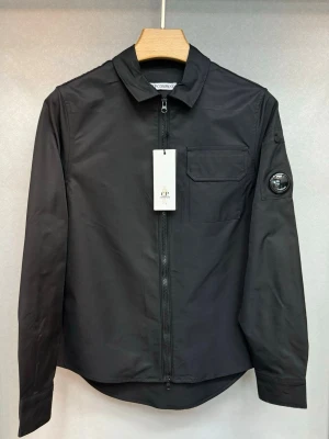 C.P. Company overshirt   - Stilren svart overshirt från C.P. Company med dragkedja framtill, klassisk krage och bröstficka. Ikonisk linsdetalj på vänster ärm. Tillverkad i ett slätt, lätt material som ger en modern look. Perfekt för lager-på-lager och streetwear.
