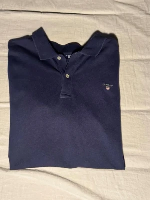 Gant piké - Storlek 170. Stilren mörkblå piké från Gant med klassisk krage och knappar framtill. Skjortan har en liten broderad logga på bröstet och är gjord i mjuk bomull för en bekväm känsla. Perfekt för dig som gillar en enkel men snygg look. Priset kan diskuteras! 