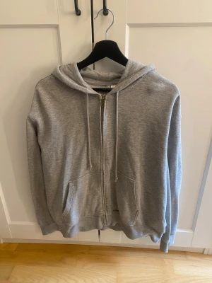 Grå zip hoodie från H&M - Klassisk grå zip hoodie från H&M med huva och dragsnören. Tillverkad i mjuk bomullsblandning och har två stora fickor framtill. Perfekt för en avslappnad och enkel stil.