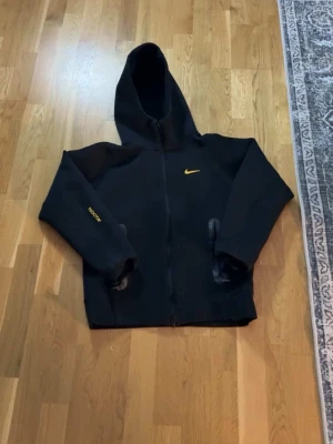 Svart Nike NOCTA hoodie med dragkedja - Säljer en svart hoodie från Nike NOCTA-kollektionen med gul logga på bröstet och NOCTA-tryck på ärmen. Hoodien har dragkedja framtill, huva och detaljer i gult på både fram- och baksida. Perfekt för dig som gillar streetwear och vill sticka ut.
