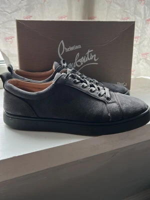 Gråa sneakers från Christian Louboutin - Stilrena gråa sneakers från Christian Louboutin i mocka med matchande grå skosnören och svart sula. Klassisk låg modell med diskret design och mjukt innerfoder i läder. Perfekta för dig som gillar exklusiva och minimalistiska skor.