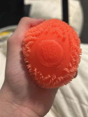 Orange Nee Doh fuzzball wonder waves - Säljer en orange stressboll från Nee Doh med mjukt gummi och taggig yta. Bollen är rund och har ett coolt mönster med vågor och små piggar som gör den extra skön att klämma på. Perfekt för dig som gillar att ha något att pilla med.