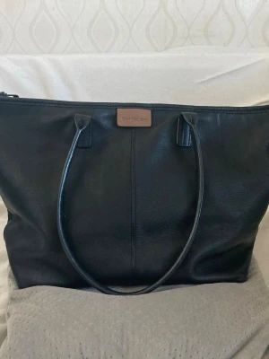 Svart toteväska från Calvin Klein Jeans - Stilren svart toteväska från Calvin Klein Jeans i äkta läder. Väskan har två långa handtag och en beige logodetalj framtill. Perfekt för dig som vill ha en rymlig och klassisk handväska med enkel design.
