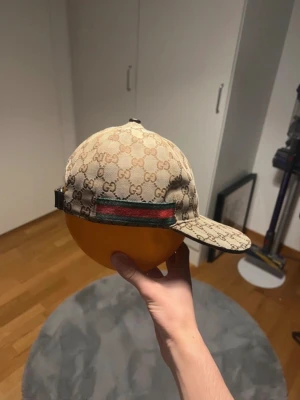 Gucci keps med GG-mönster - Säljer en beige keps från Gucci med klassiskt GG-monogram över hela kepsen. Kepsen har en böjd skärm och justerbar rem bak. På sidan finns den ikoniska gröna och röda randen. Perfekt för dig som vill ha en lyxig och trendig accessoar.