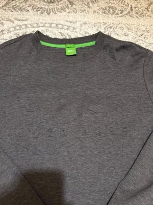 Mörkgrå sweatshirt från Hugo Boss - Snygg mörkgrå sweatshirt från Hugo Boss med diskret broderad logga på bröstet. Tröjan har rund halsringning, långa ärmar och ribbade muddar. Tillverkad i mjukt bomullsmaterial och har en grön detalj på insidan av kragen.