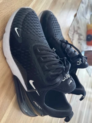 Nike Air Max 270 svarta sneakers Stl.37 - Nike Air Max 270 sneakers i svart med vita detaljer och stor synlig Air-enhet i hälen. Ovandelen är i mesh för bra ventilation och skon har klassiska Nike-loggor på sidan och tån. Snygg och sportig design som passar till streetwear.