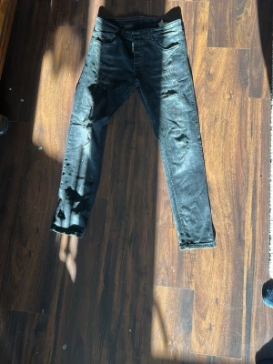 Svarta dsquared2 jeans  - Säljer ett par svarta skinny jeans med coola slitningar och färgstänk. Jeansen har klassisk femficksdesign och smal passform. Perfekta för dig som gillar en edgy och trendig stil.