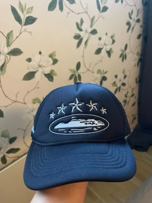 Svart keps  - Säljer en svart truckerkeps med mesh på sidorna och broderade stjärnor samt ett UFO-motiv framtill. Kepsen har böjd skärm och justerbar baksida. Perfekt för dig som gillar streetwear och unika detaljer.