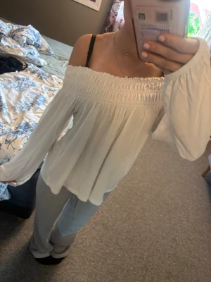Vit off shoulder blus med vida ärmar - Säljer en vit off shoulder-blus med smock upptill och långa, vida ärmar. Blusen har en luftig och flowy passform, perfekt för en avslappnad och trendig look. Materialet känns mjukt och lätt mot huden.