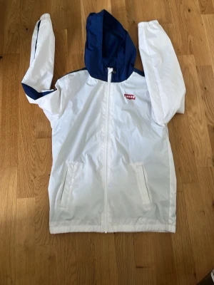 Vit och blå vindjacka från Levi's - Snygg vit vindjacka från Levi's med blå huva och blå detaljer på ärmarna. Jackan har dragkedja framtill, två fickor och stor Levi's-logga på ryggen. Perfekt för blåsiga dagar och streetwear-stil.