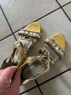 Guldiga sandaler med nitar storlek 37 - Säljer ett par glansiga guldiga sandaler med breda remmar och coola silverfärgade nitar. Sandalerna har öppen tå och en liten klack, perfekta för att sticka ut. Materialet är syntet med metallic-finish och dekorationer i plast/metall.