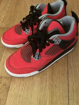 Röda Air Jordan 4 sneakers - Snygga Air Jordan 4 sneakers i rött med svarta och grå detaljer. Skorna har klassisk meshpanel på sidorna, svart snörning och vit sula. Ikoniska Jumpman-loggan syns på hälen. Perfekta för dig som gillar streetwear och sportig stil.