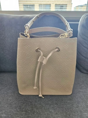 Louis bucket bag - Stilren beige handväska från Louis i bucket-modell med präglat monogrammönster. Väskan har både axelrem och handtag i läder, samt guldfärgade metalldetaljer och dragskostängning. Perfekt för dig som vill ha en lyxig och trendig accessoar.