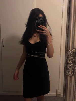 Svart klänning med pärldetaljer - Svart klänning med v-ringad hals och pärldetaljer runt midjan. Klänningen är ärmlös och har en elegant, figurnära passform som framhäver siluetten. Perfekt för dig som vill ha en stilren look med lite extra bling.