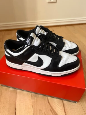 Nike Dunk Low svart/vit sneakers - Nike Dunk Low sneakers i svart och vitt läder med klassisk Swoosh på sidan. Lågt skaft, svart snörning och vit mellansula. Tidlös design med perforerad tåbox och Nike-logga på plös och häl. Perfekta för dig som gillar streetwear och stilrena färger.