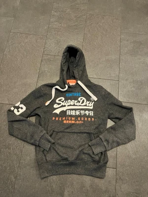 Mörkgrå Superdry hoodie med tryck - Snygg mörkgrå hoodie från Superdry med stor vit logga och japanskt tryck på bröstet. Har huva med vita dragsnören, magficka och tryckt siffra på ärmen. Perfekt för en avslappnad streetstyle-look.
