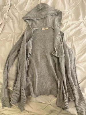 Grå stickad hoodie från Hollister - Mysig grå stickad hoodie från Hollister med långa ärmar och ribbade muddar. 