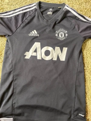 Manchester United träningströja Adidas S - Mörkgrå Manchester United träningströja från Adidas med vita detaljer och logga. V-ringad modell med ClimaCool-material som andas. Stora AON-trycket på bröstet och klassiska Adidas-ränder på axlarna. Perfekt för fotbollsträning eller gym.