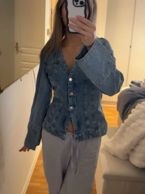 Figursydd jeans tröja med vida ärmar - Snygg figursydd jeans tröja i ljusblå denim med markerade sömmar och vida ärmar. Tröjan har knäppning framtill med silvriga knappar och en lätt v-ringad krage. Endast använd en gång. 