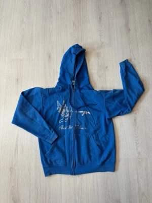 Rhinestone Hoodie - Blå hoodie med dragkedja och huva. Framtill finns ett tryck med en fågel och ett gevär'. Hoodien har fickor på sidorna och justerbara snören i huvan. Perfekt för en avslappnad streetwear-look.