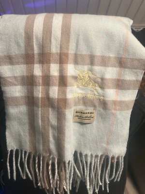 Burberry halsduk i ljusblå kashmir - Snygg ljusblå halsduk från Burberry i 100% kashmir med klassiskt rutmönster i beige, vitt och ljusrosa. Broderad Burberry-logga och fransar i kanterna ger en exklusiv känsla. Perfekt accessoar för att lyfta din outfit.