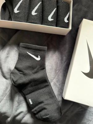 Svarta Nike strumpor med vit logo - Säljer ett paket svarta Nike strumpor med den klassiska vita swoosh-loggan. Strumporna är ribbade och har en sportig look, perfekta för dig som gillar streetwear. Kommer i originalförpackning med fem par. Materialet är mjukt och bekvämt.