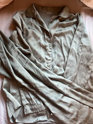 Ljusgrön satinpyjamas med skjortkrage - Snygg ljusgrön pyjamas i satin med klassisk skjortkrage och knäppning framtill. Långa ärmar och fickor framtill ger en chill och stilren look. Perfekt för mysiga hemmakvällar.