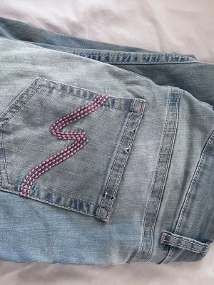 Ljusblå jeans med rosa söm - Säljer ett par ljusblå jeans med en unik rosa zigzag-söm på bakfickan. Jeansen har klassisk femficksdesign och detaljer i rosa vid bälteshällor och knapphål. Perfekta för dig som vill ha något extra i din denimlook.