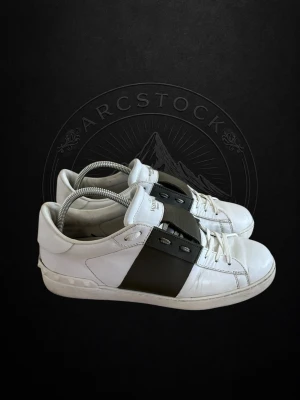 Valentino Opens - Snygga vita sneakers från Valentino Garavani med bred svart läderdetalj över mitten och ikoniska nitar på hälen. Skorna har klassisk låg profil, vita snören och är tillverkade i läder för en lyxig känsla.