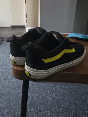 Svarta Vans sneakers med gul detalj - Använd 3 ggr. Svarta Vans sneakers i mocka med den klassiska gula sidoremsan och vit sula. Skorna har lågt skaft och snörning framtill. Perfekta för en streetwear-look och passar till det mesta. Tidlös design med tydlig Vans-logga på häl och plös. Skriv för mer info 