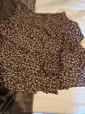 Brun blommig volangkjol från Lindex - Supersöt brun kortkjol från Lindex med vita små blommor och volangdetaljer. Kjolen har resår i midjan och är perfekt för dig som gillar en avslappnad och trendig stil. Lätt och luftig, passar perfekt till varmare dagar.