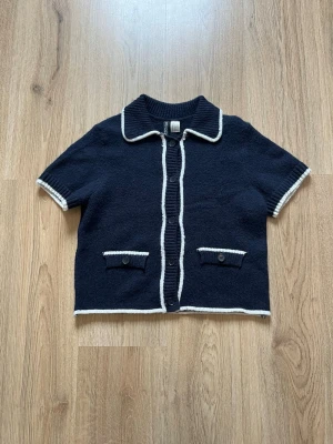 Marinblå kofta med vita detaljer - Kortärmad marinblå kofta med vita kanter och dekorativa fickor framtill. Koftan har knappar längs hela framsidan och en klassisk krage. Perfekt för en stilren och retroinspirerad look.