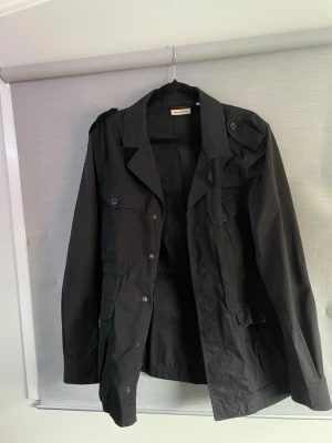 Fieldjacka  - Stilren svart fieldjacket från dkny med klassisk krage, knäppning framtill och flera fickor med lock. Jackan har axelklaffar och är tillverkad i ett lätt material som passar perfekt för blåsiga dagar.