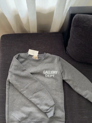 Grå sweatshirt från Gallery Dept. - Snygg grå sweatshirt från Gallery Dept. med vit logga tryckt på bröstet och stor text på ryggen. Klassisk rund halsringning och ribbade muddar vid ärmslut och nederkant. Perfekt för en avslappnad streetwear-look.