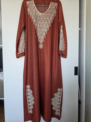 Lång brun klänning med broderi - Långklänning i varm brun ton med vackra vita broderier längs halsringning, ärmar och nedtill. Klänningen har lång ärm och ett löst, bekvämt fall. Perfekt för dig som gillar unika detaljer och bohemisk stil.